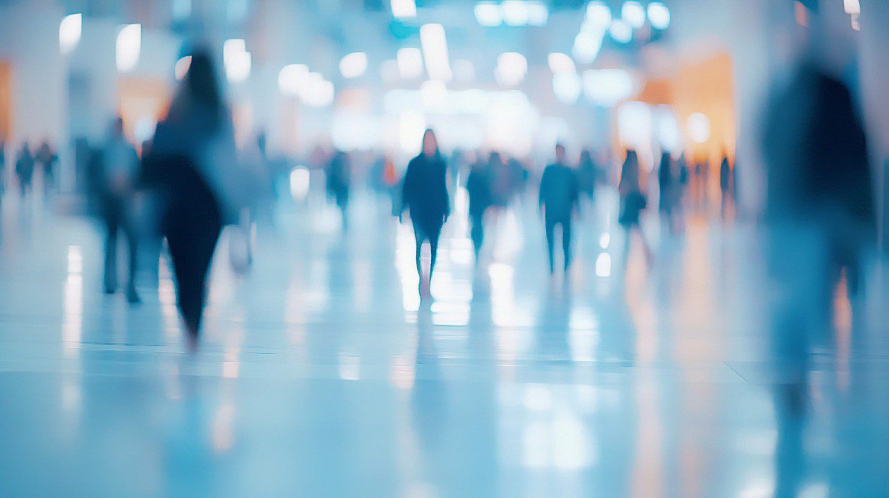 Blurred silhouettes of people walking through a brightly lit, modern exhibition space with soft glowing lights and motion blur effect.