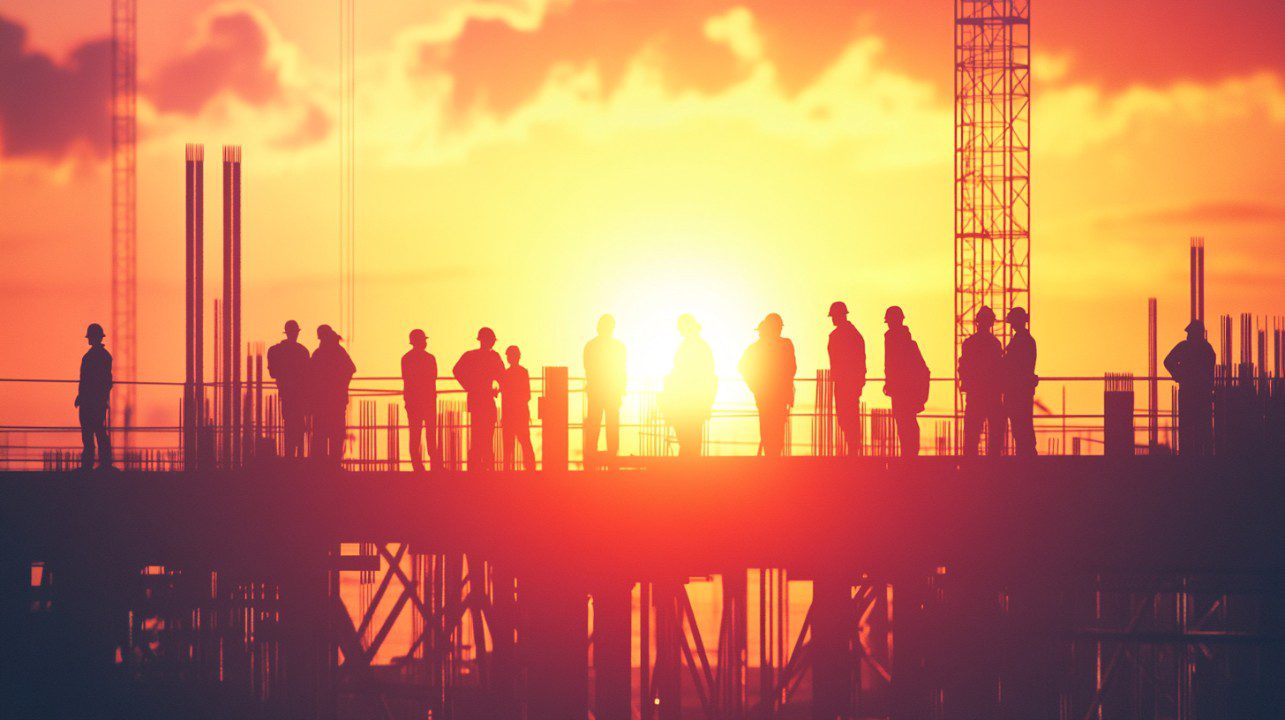 Silhouette of a group of construction workers standing on a job site during sunset, evoking themes of teamwork and industry.