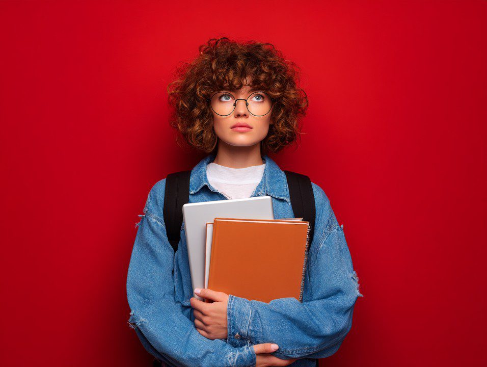 Curly-haired young student wearing glasses and a denim jacket holding notebooks while standing against a vivid red wall.