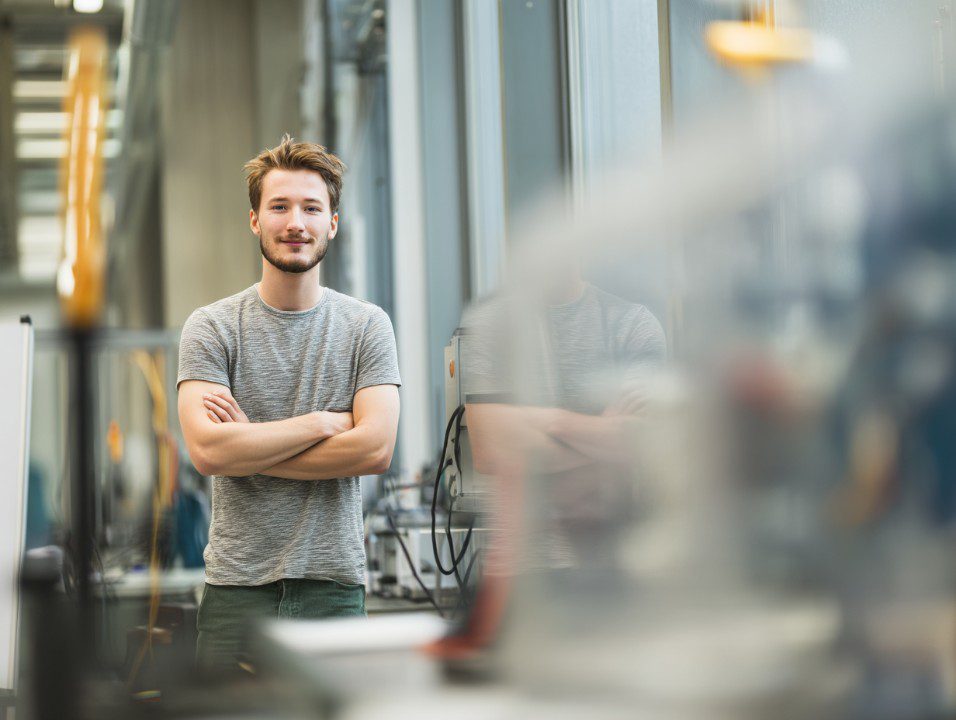 Young engineering student standing confidently with arms crossed in a modern university laboratory filled with technical equipment.