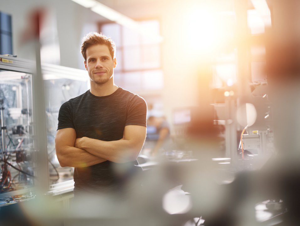 Confident young engineering student with arms crossed in a sunlit university lab, surrounded by technical equipment and machinery.