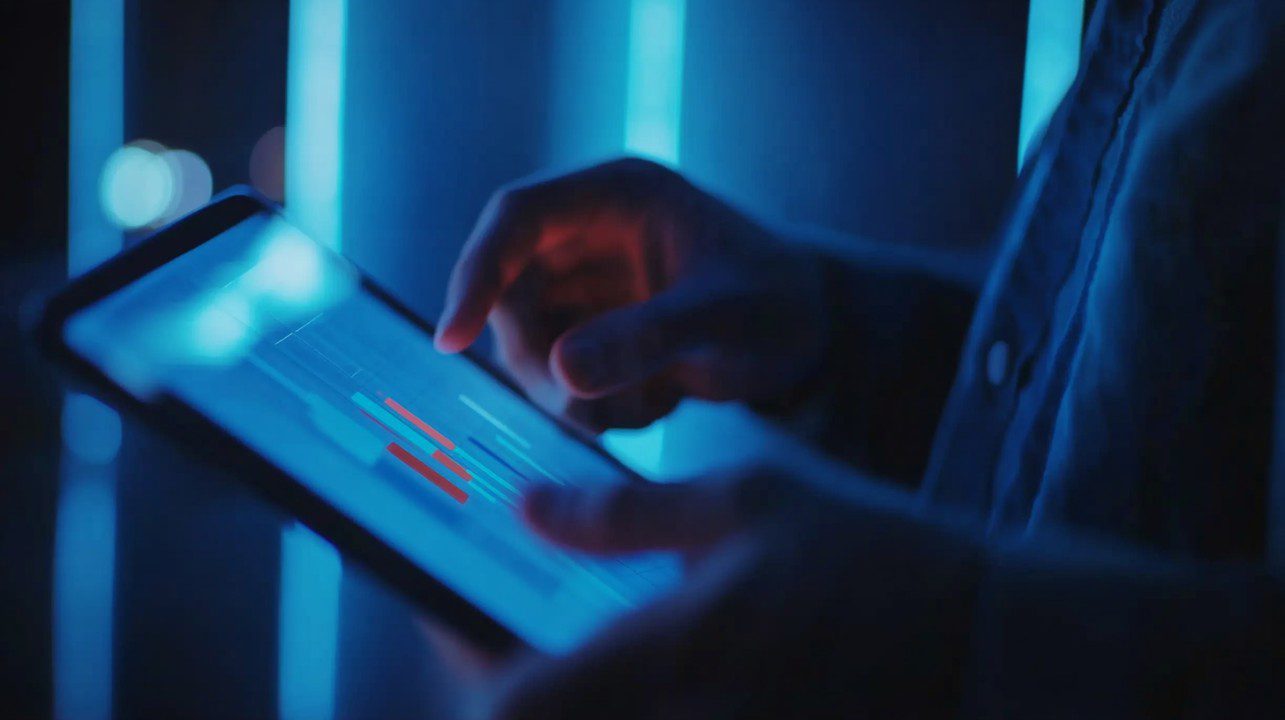 Close-up of person using a tablet with financial charts on screen in a modern, dark-lit fintech environment.
