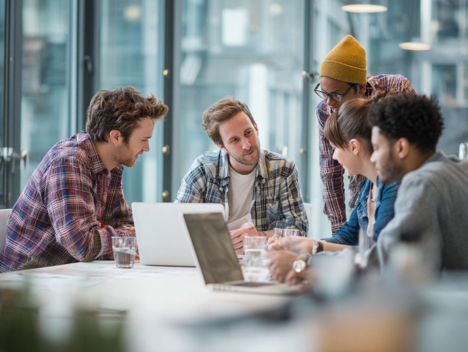 Group of young professionals collaborating around a desk in a modern glass office, showcasing teamwork and creative discussion.
