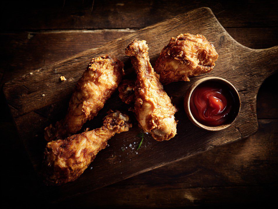Golden crispy fried chicken drumsticks on a rustic wooden board with a small bowl of ketchup, showcasing rich texture and flavor.