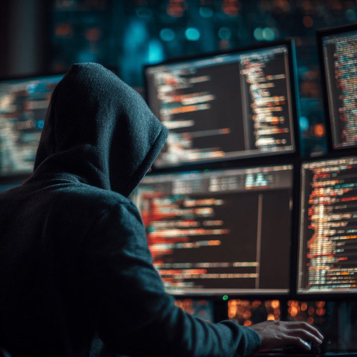Hooded hacker in a dark room working on multiple coding screens, representing cybersecurity threats and hacking activity.