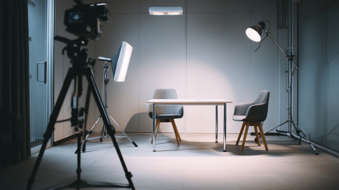 Professional interview setup featuring studio lighting, two chairs, a camera tripod, and a modern minimalist background.