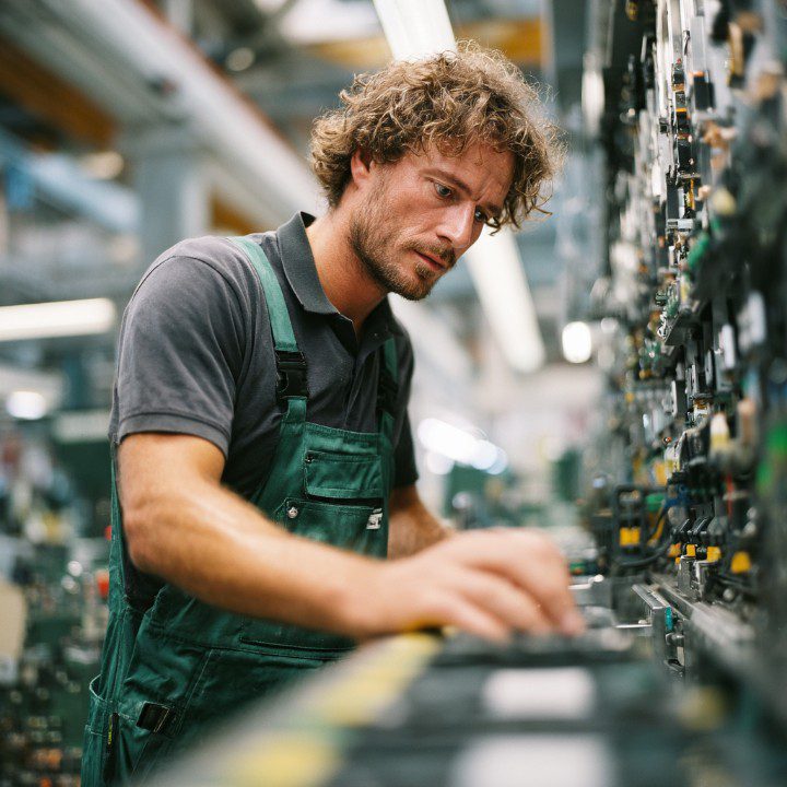 Focused mechatronics technician in green overalls adjusting an electrical control panel in a modern industrial facility.