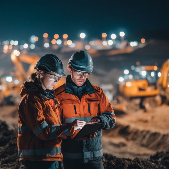 Two engineers in safety gear using a tablet at a nighttime construction site, surrounded by illuminated heavy machinery and equipment.