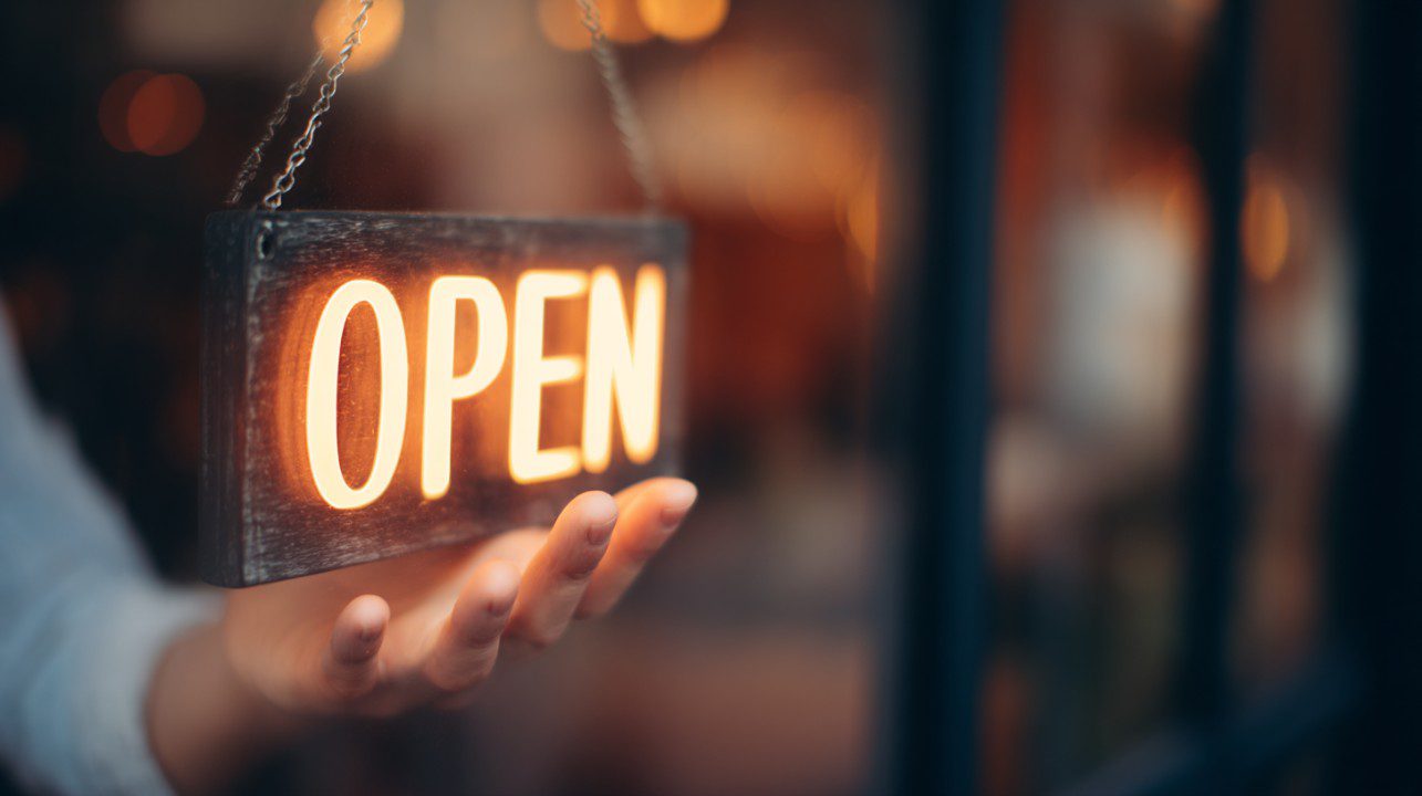A glowing "OPEN" sign being held in front of a window, symbolizing an open store or welcoming business atmosphere.
