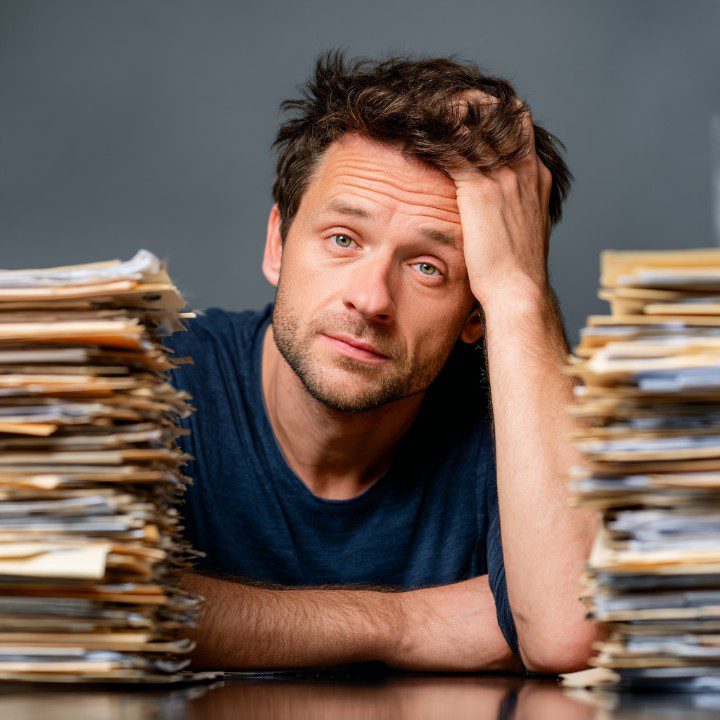 A stressed man in a dark blue shirt sitting between two tall piles of paperwork, resting his head in frustration—symbolizing burnout or work overload.