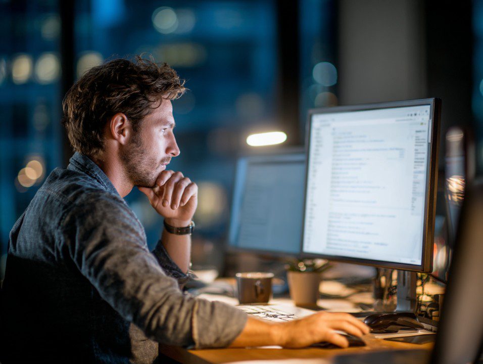Man concentrating on a desktop computer late at night in a modern office, surrounded by monitors and dim lighting.
