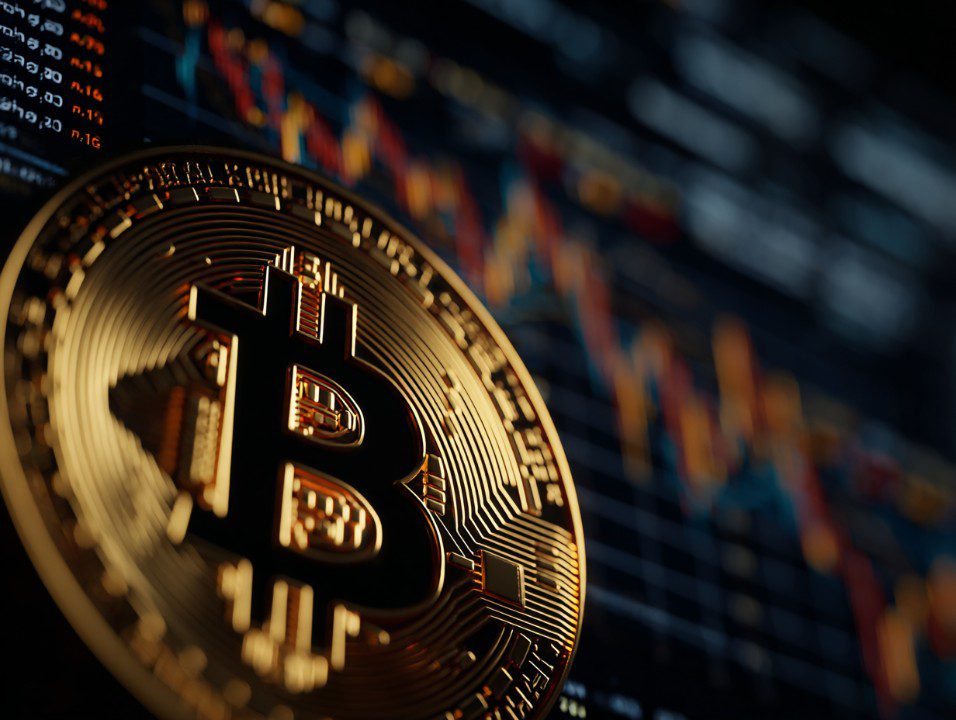 A close-up of a golden Bitcoin coin with colorful cryptocurrency trading charts in the background, symbolizing digital finance.
