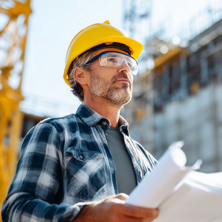 Construction worker wearing a yellow hard hat and protective glasses holding blueprints while overseeing an active building site.