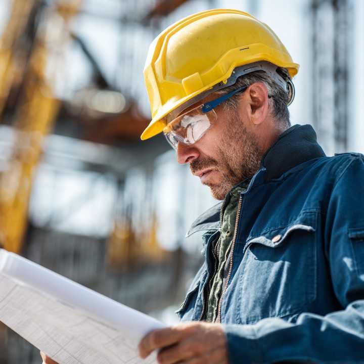 Construction worker wearing a yellow hard hat and glasses, reviewing blueprints at a job site with structural steel in the background.