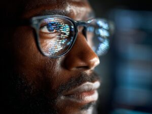 Close-up of a man’s glasses reflecting lines of computer code, representing cybersecurity, programming, and digital vigilance.