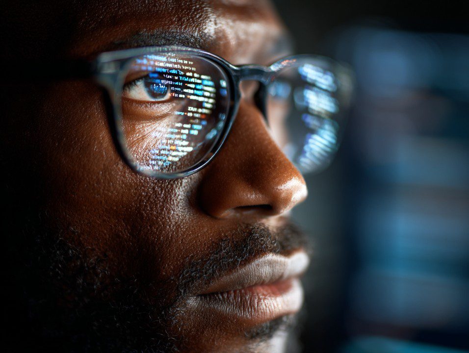 Close-up of a man’s glasses reflecting lines of computer code, representing cybersecurity, programming, and digital vigilance.