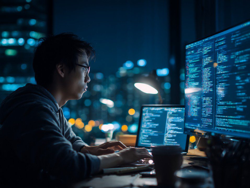 Focused programmer working late at night in city office with multiple monitors displaying lines of code.