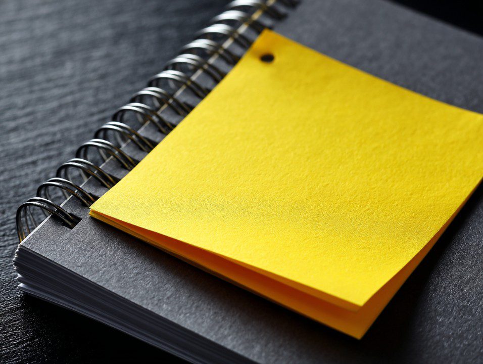 Close-up of a yellow sticky note attached to a gray spiral notebook, symbolizing reminders, organization, or productivity.