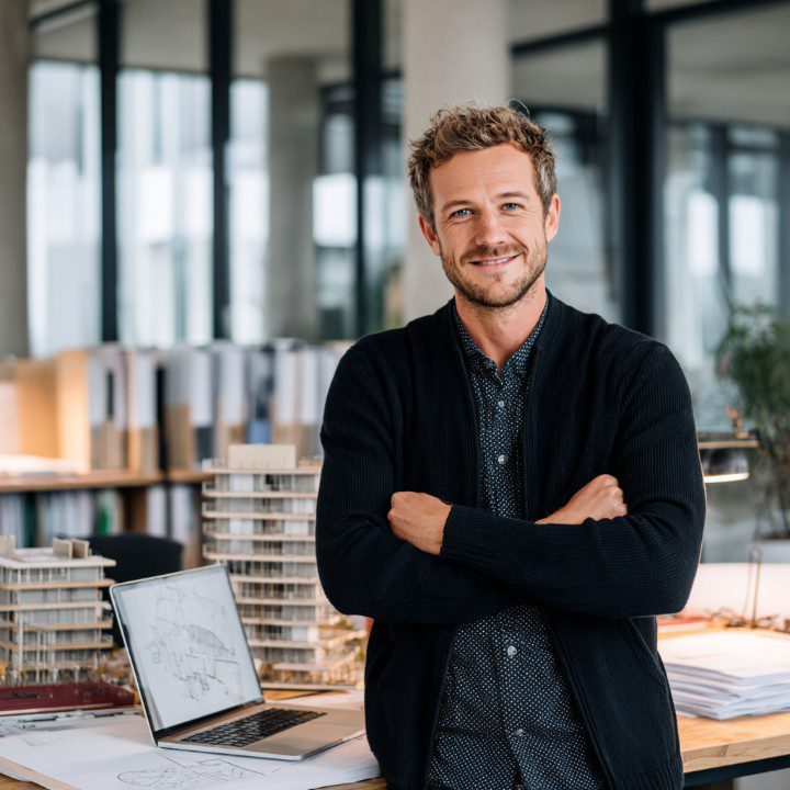 A smiling architect stands in a modern office with arms crossed, surrounded by building models, blueprints, and a laptop showing architectural drawings.