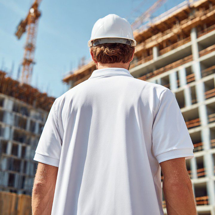 Construction worker wearing a white hard hat, viewed from the back, overseeing a building site under construction.