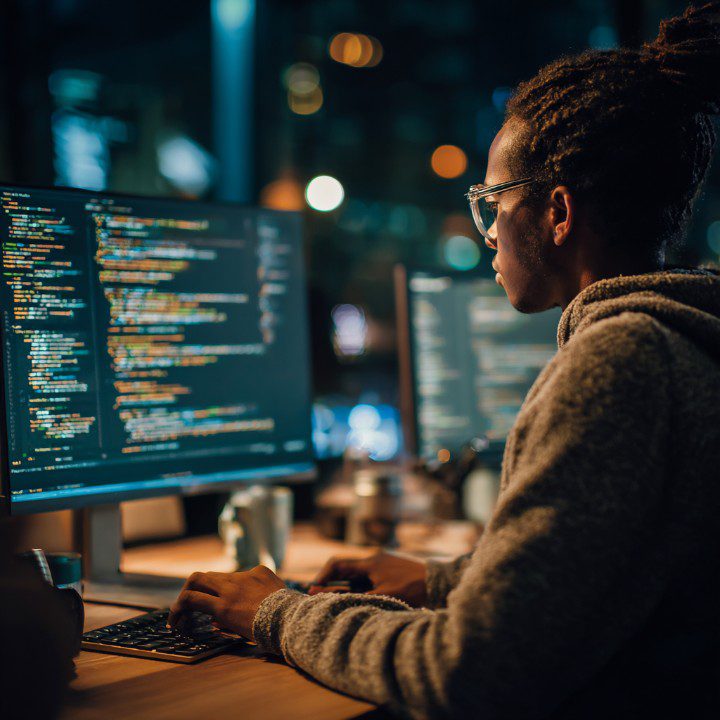 A programmer in glasses works at night on dual monitors filled with colorful code in a modern low-light workspace.