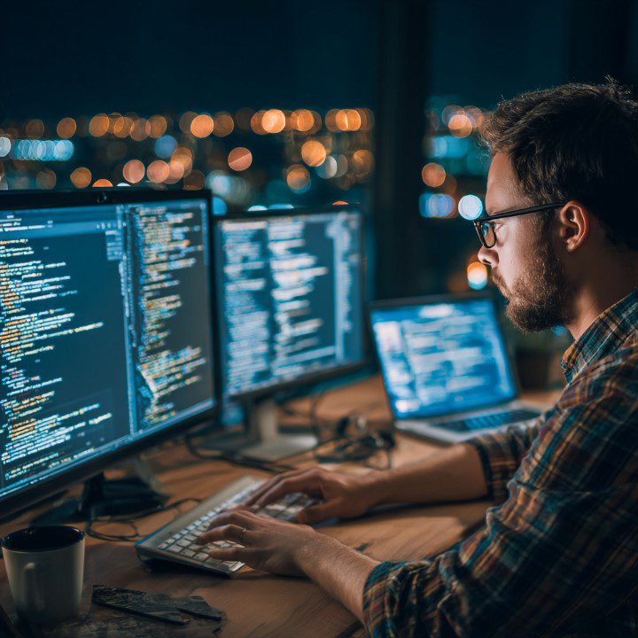 Programmer coding at night on multiple monitors with city lights in the background.