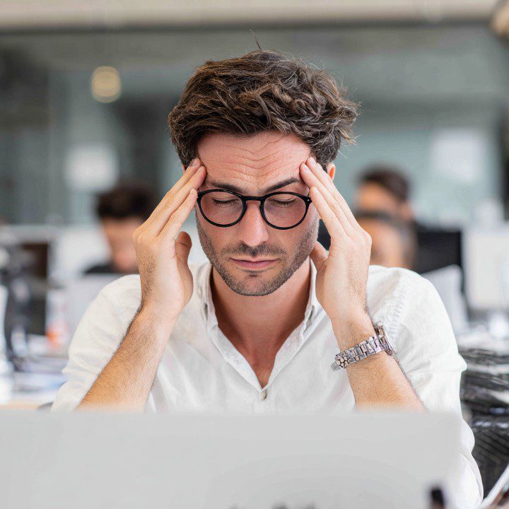 Man in glasses holding his temples in stress while working in an office environment.