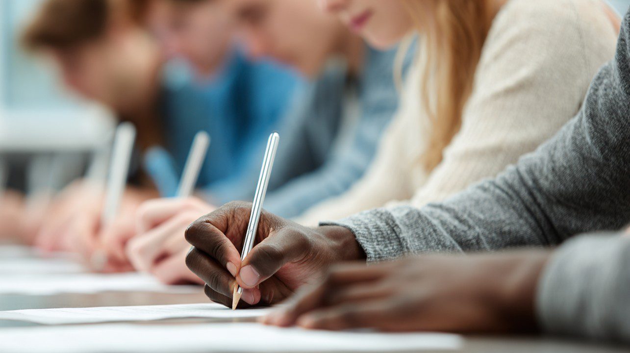 Close-up of students' hands writing with pens during an exam, with blurred background of classmates.