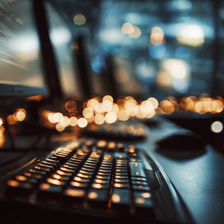 Close-up of a backlit keyboard with bokeh lighting and dual computer monitors in a tech-focused workspace.