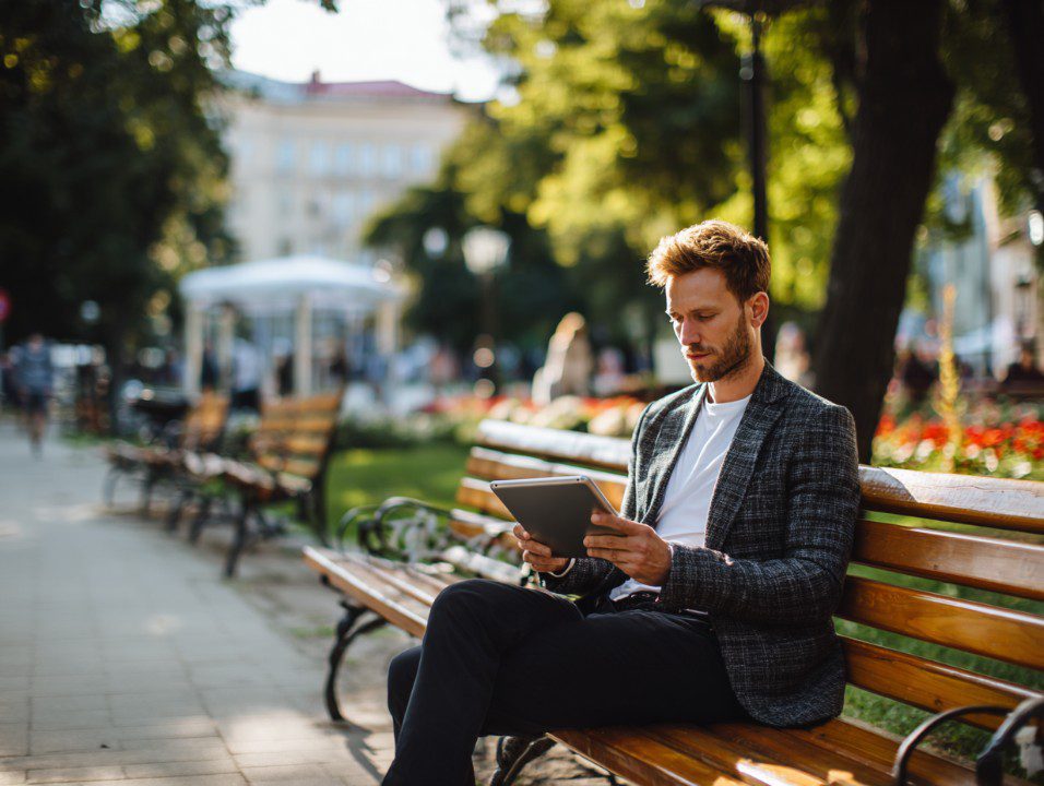 Stylish man sitting on a city park bench using a tablet, representing remote work, digital lifestyle, and modern productivity.